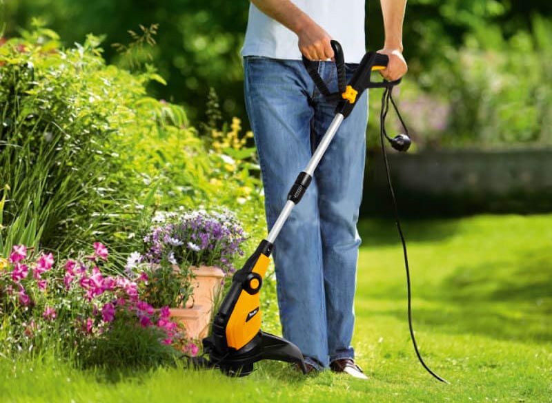 Підстригання трави електричним тримером біля клумби